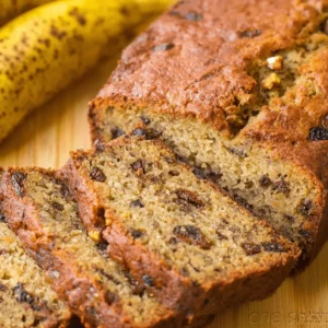 A warm, golden-brown loaf of homemade pumpkin banana bread with fresh slices and ripe bananas on a wooden board.